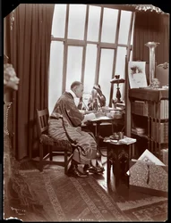 Will R. Barnes i en mantel, sittande vid sitt skrivbord i sin studio, 1903 (silvergelatintryck)